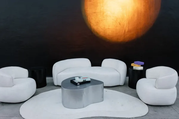 Lounge seating area with a white curved sofa and white lounge chairs around a marble coffee table, set before a painted mural wall
