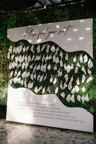 Seating chart display with a wedding seating chart sign and hanging escort cards tied to a white board against a greenery wall backdrop