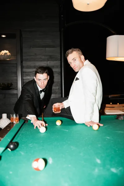 Groomsmen portrait with groomsmen playing pool, groom in white dinner jacket and bow tie holding whiskey by a pool table in a dark lounge