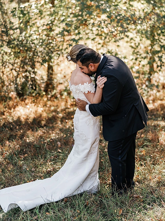 Wedding couple portrait of bride and groom hug in a sunlit woodland, her off-the-shoulder floral appliqué gown and train on fallen leaves