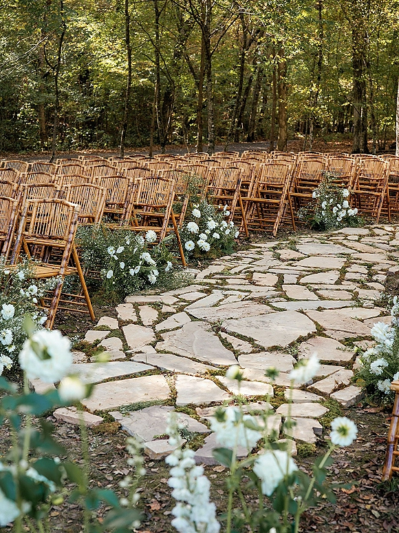 Outdoor ceremony setup with woodland wedding ceremony rows of wood chairs lining a stone aisle, white flowers and greenery in a forest clearing