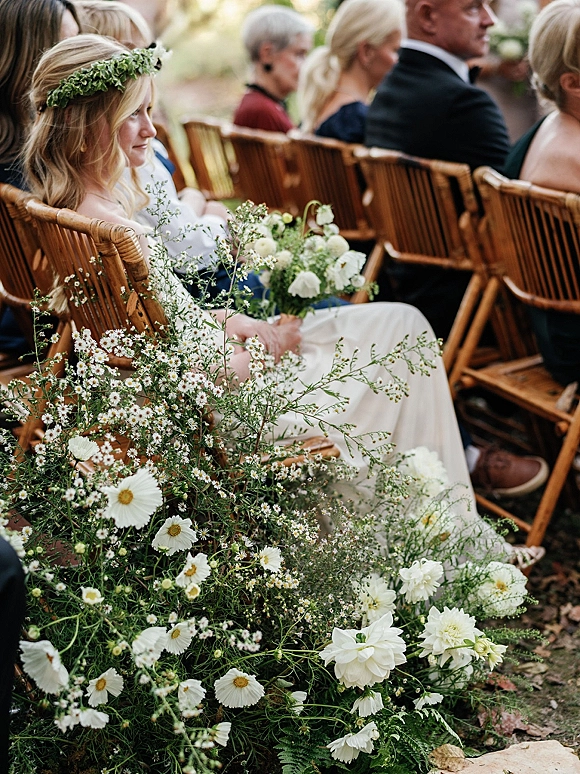Ceremony guests seated on wooden folding chairs with aisle florals, one wearing a flower crown in an outdoor foliage setting