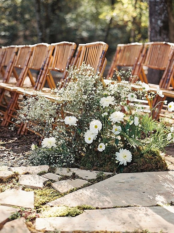 Ceremony aisle decor with outdoor ceremony aisle flowers, white daisies and baby’s breath on moss beside wood chairs along a stone path in forest light