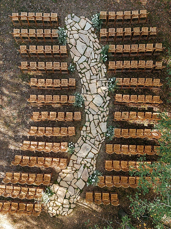 Ceremony aisle design with a stone aisle runner between wood chairs, lined with white flowers and greenery beneath trees