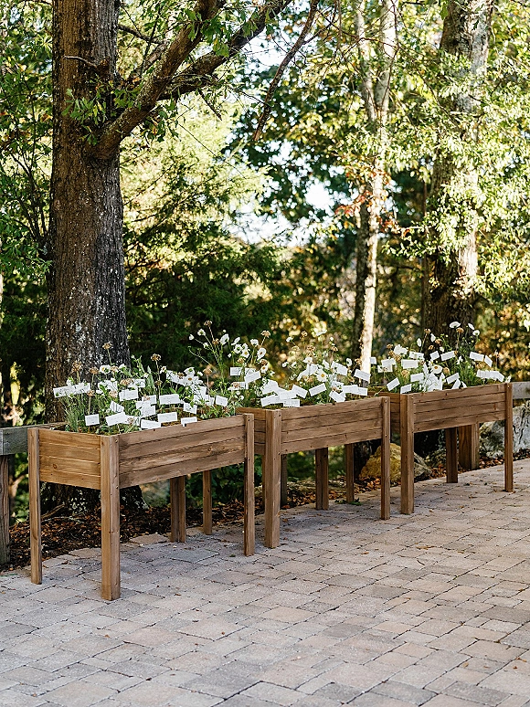 Wedding escort cards in a wood planter escort display with wildflowers and greenery on a stone patio surrounded by garden trees