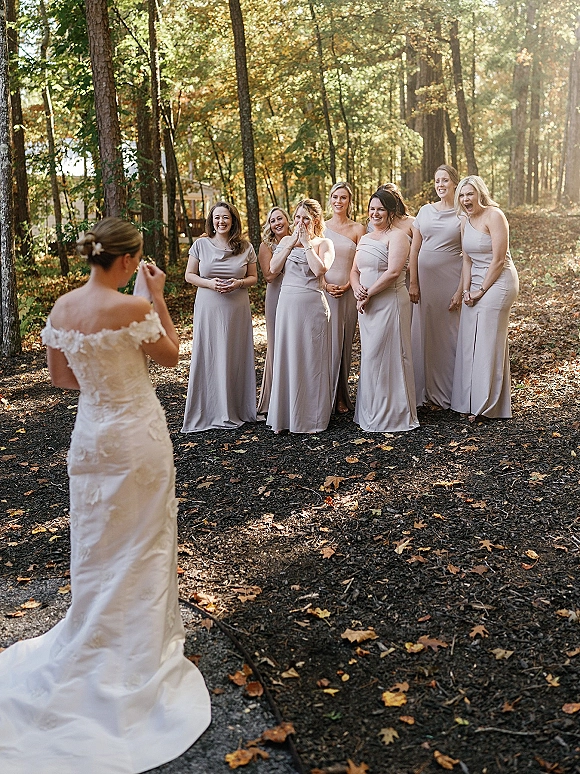 Bridesmaid reveal moment as the bride in an off-shoulder lace gown with long train walks up a sunlit woodland path in fall leaves