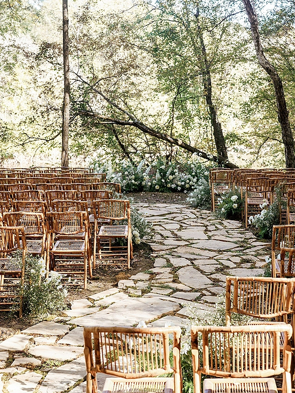 Outdoor ceremony setup with wooden chairs and a stone aisle lined with white florals and greenery beneath sunlit woodland trees
