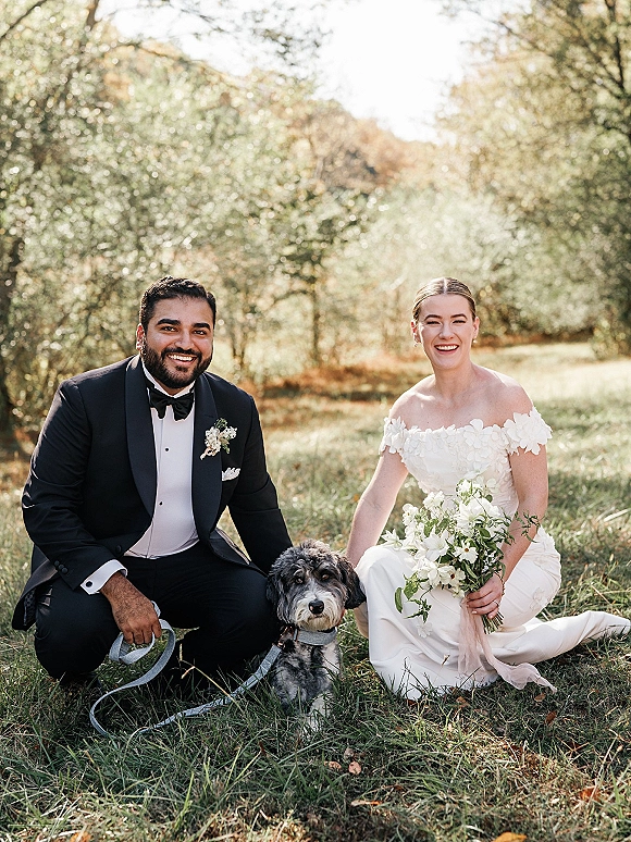 Couple portrait of bride and groom with dog on a sunlit park lawn, bride holding a white and green bouquet in an off-shoulder gown