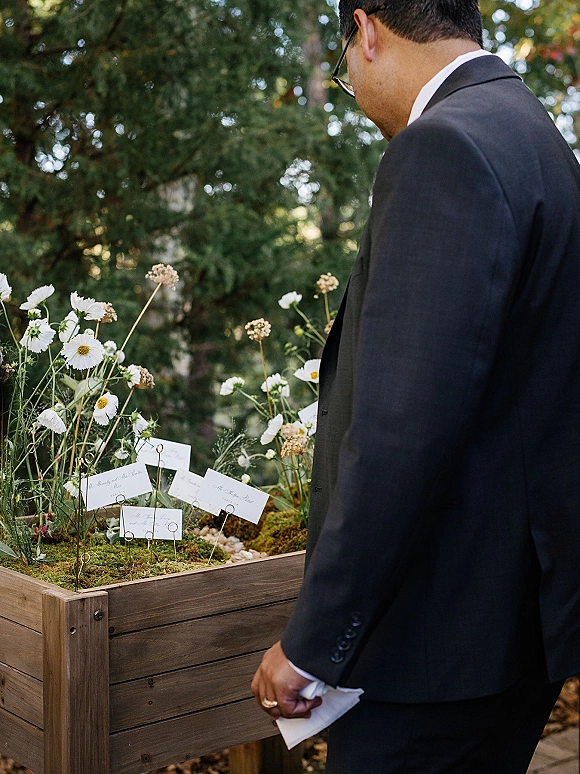 Wedding escort cards in a wooden planter box with wire holders, moss and wildflowers, as a groom in glasses reads beside trees
