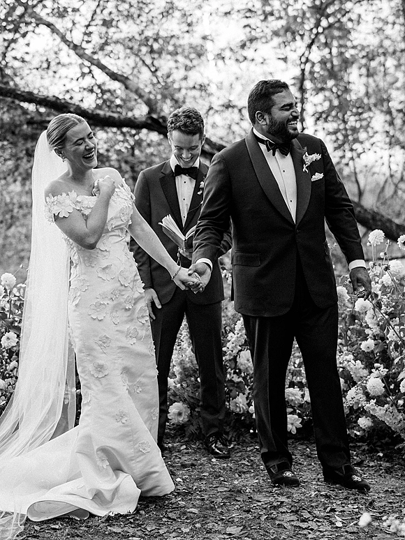Wedding recessional as bride and groom walk hand in hand, laughing in a garden aisle, her long veil flowing beside his tuxedo