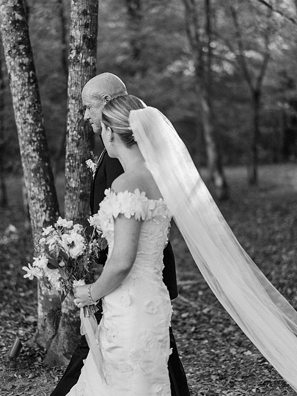 Wedding processional as a bride walking down aisle with bouquet and long veil beside her father on a woodland path with fallen leaves