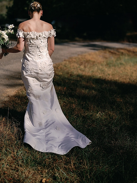 Bridal portrait of a bride in a back view wedding dress, holding a white bouquet and walking along a shaded tree-lined path