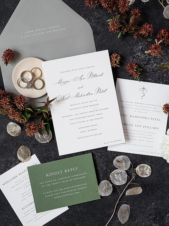 Invitation suite wedding invitation flat lay with calligraphy cards, envelopes, wax seal, rings in dish, and pressed leaves on dark stone surface