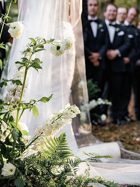 Ceremony moment as bride from behind walks to altar, long veil and gown train flowing past white floral and greenery aisle in garden