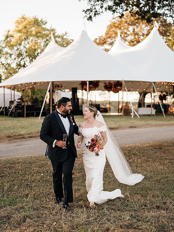 Wedding couple portrait of bride and groom walking on lawn, holding champagne flutes; her veil and burgundy bouquet by lit white tent background