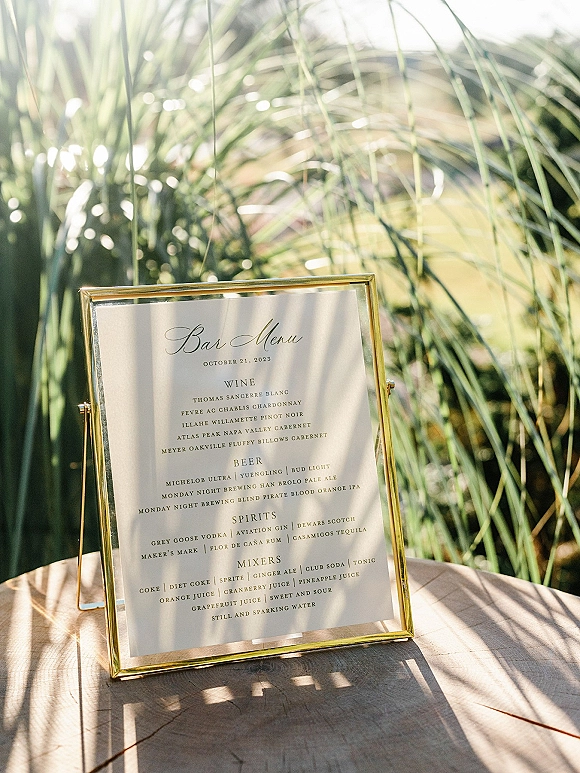 Wedding bar menu in a gold frame, a signature cocktail menu sign on an acrylic stand atop a wooden table in sunlit greenery