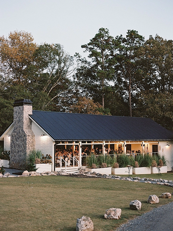 Wedding reception venue with a white barn exterior, covered porch string lights, and a stone chimney beside a lawn and trees under open sky