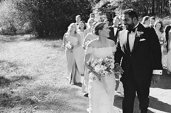 Wedding party portrait of bride and groom walking with bridesmaids and groomsmen on a sunlit lawn, bride holding bouquet