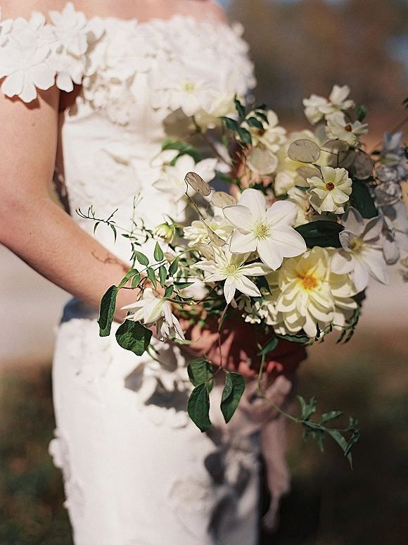 Bridal bouquet of white wedding bouquet blooms and greenery against a ruffled bridal gown, with soft outdoor foliage blurred behind