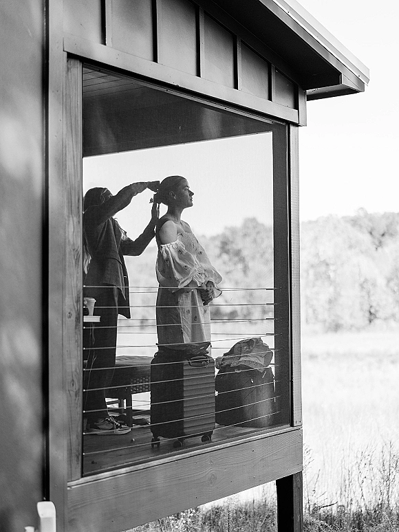 Bridal hair styling as a hairdresser shapes a bride hair bun with pins on a modern cabin porch, luggage by bench, meadow beyond window