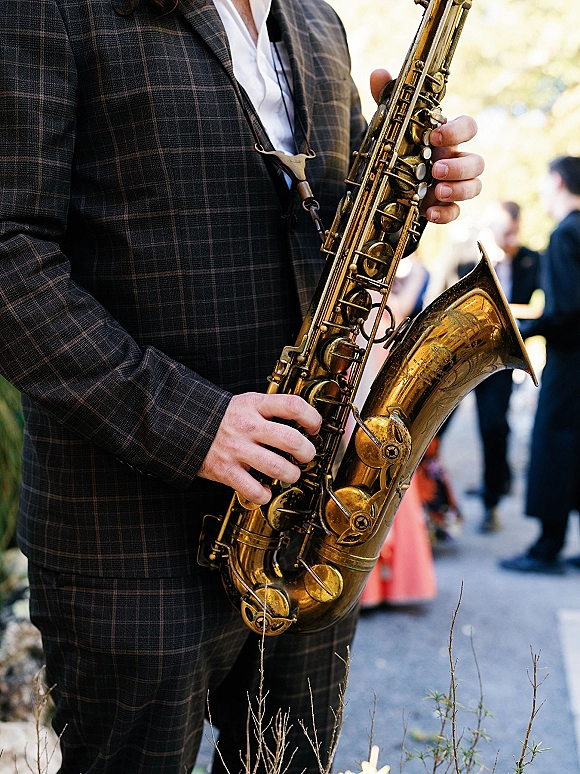 Wedding saxophonist playing live wedding music in a plaid suit, saxophone on neck strap, on an outdoor walkway with greenery and guests