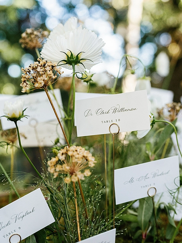 Wedding place cards in gold card holders with wildflower stems, forming an escort card display against soft garden bokeh and trees