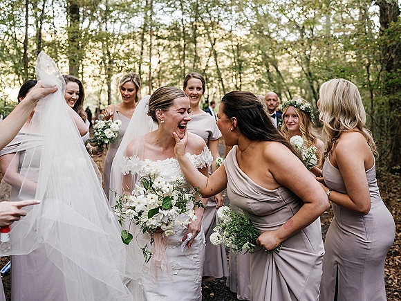 Bride with bridesmaids laughing as they adjust her wedding veil on a forest path, holding white bouquets in blush dresses