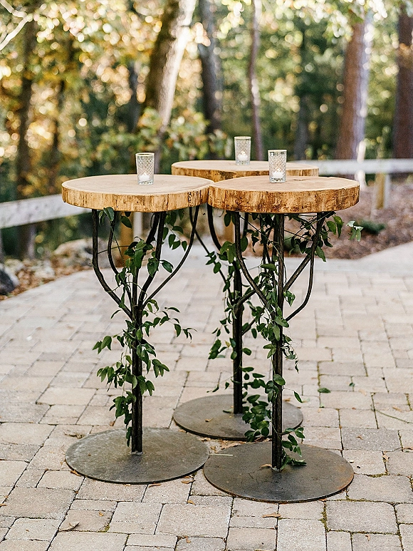 Cocktail table decor with wood slab cocktail tables on metal pedestals, greenery garland and glass votive candles on a garden patio