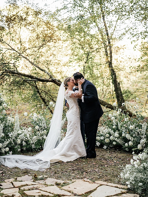 Wedding kiss portrait of bride and groom kissing on a stone garden path, her long veil and lace dress trailing beside aisle flowers