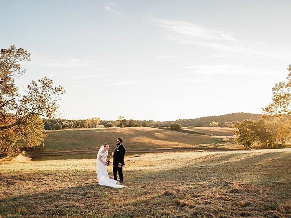 Couple portrait in sunset wedding photos, bride in lace dress and veil holding bouquet beside groom in tuxedo in open field with hills