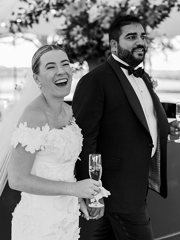 Wedding reception moment as bride laughs with groom during a champagne toast, holding flutes under hanging greenery with blurred guests behind
