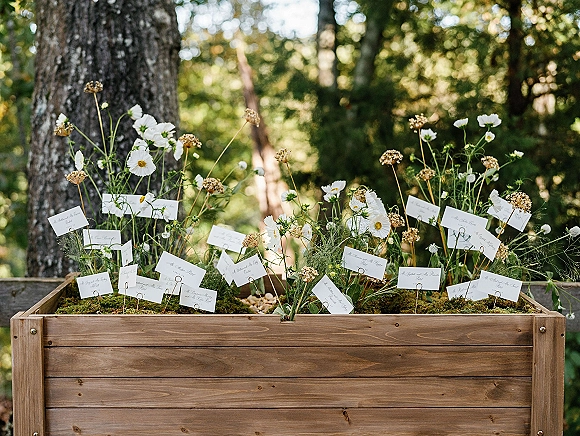 Escort card display with wedding seating cards on wire holders in a wood planter box, tucked into moss and wildflowers in a garden setting