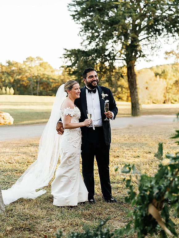 Couple portrait of bride and groom holding champagne flutes, her veil flowing in golden hour on an outdoor lawn with trees