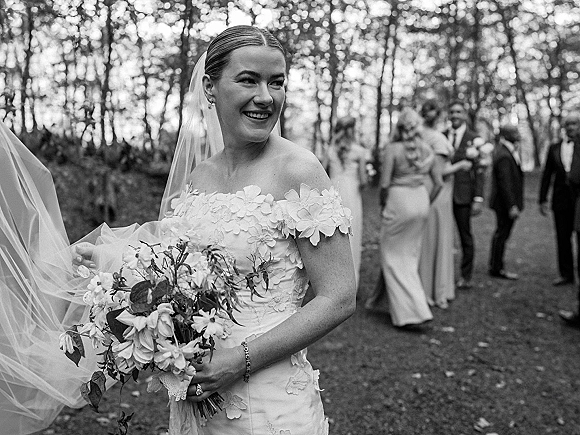 Bridal portrait in black and white of a smiling bride holding a bouquet, veil blowing softly, with wedding party blurred among trees