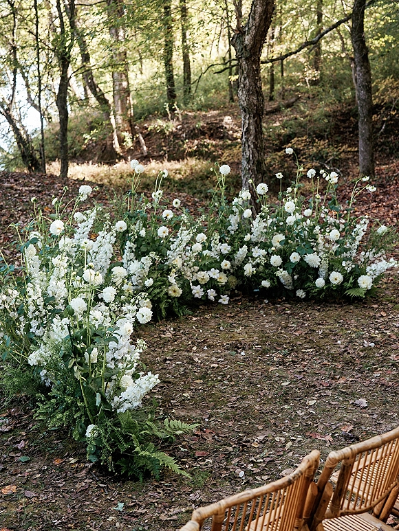 Ceremony aisle flowers in grounded aisle florals of white blooms, ferns, and greenery lining bamboo chairs in a sunlit woodland clearing