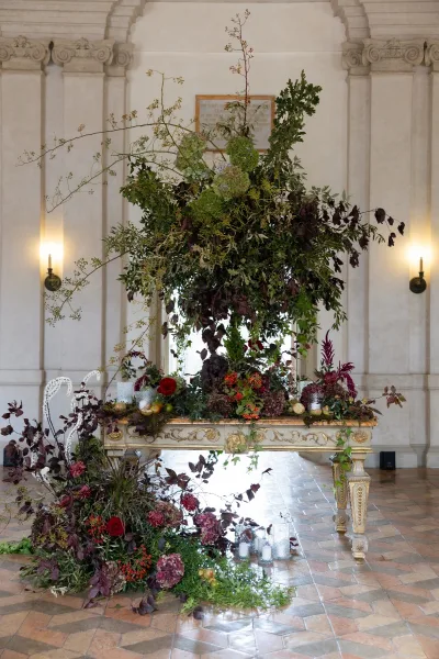 Wedding floral installation with lush greenery, hydrangeas, and red roses cascading over an ornate table with glass votive candles in a columned hall