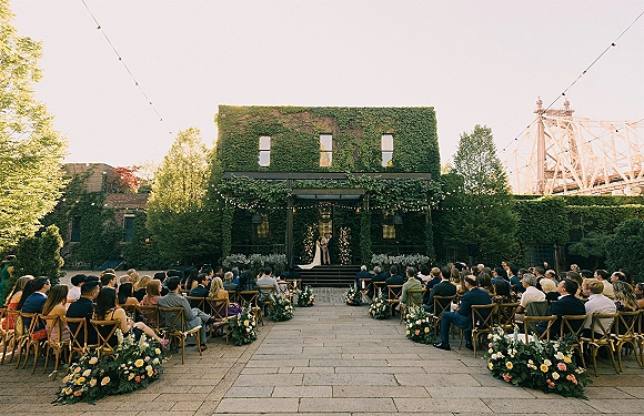 Outdoor ceremony setup with courtyard wedding ceremony chairs lining a floral aisle, string lights overhead, and an ivy-covered building backdrop
