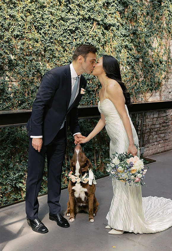 Wedding kiss portrait of bride and groom kissing while holding hands, bride with bouquet beside dog in flower collar by ivy brick wall