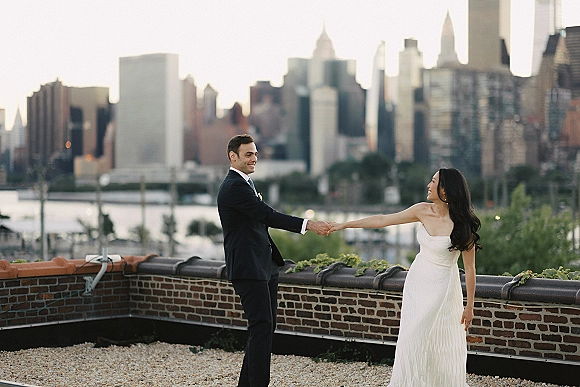 Couple portrait of bride in a strapless wedding dress and groom in suit holding hands on a rooftop with city skyline and river behind them