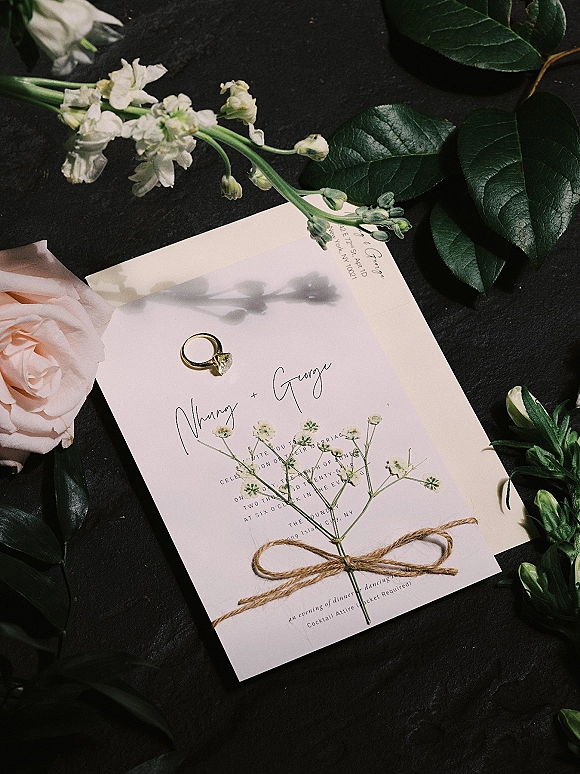 Invitation suite wedding invitation flat lay with calligraphy card, envelope, oval diamond ring, twine bow and white florals on dark fabric