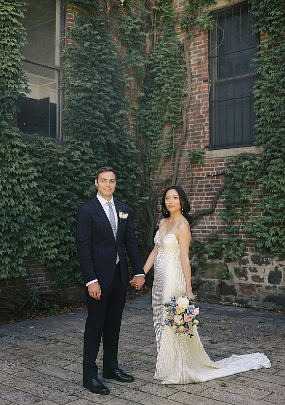 Couple portrait of bride and groom holding hands, her pastel bouquet and strapless gown against an ivy-covered brick wall courtyard.