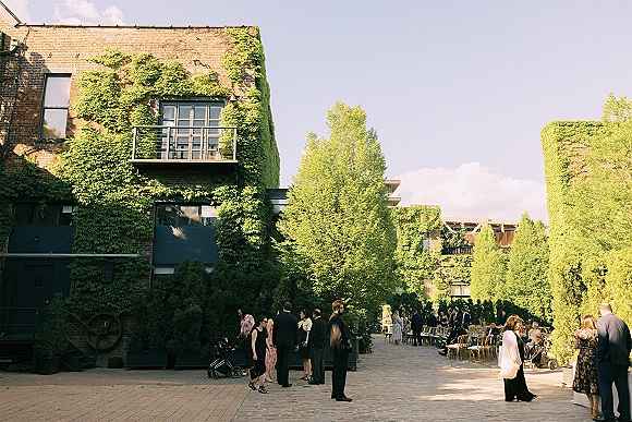 Outdoor wedding reception with wood chairs and banquet tables, floral arrangements and cocktail tables in an ivy-covered brick courtyard