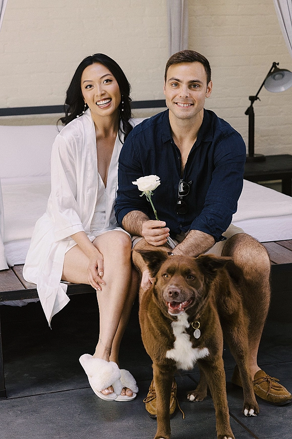 Couple portrait with dog sitting on a bed in a modern bedroom, bride in white satin robe and slippers with drop earrings by brick wall