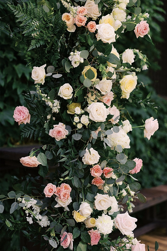 Wedding floral arrangement of blush and ivory roses with eucalyptus accent and ferns on a ceremony pillar against garden greenery and railing