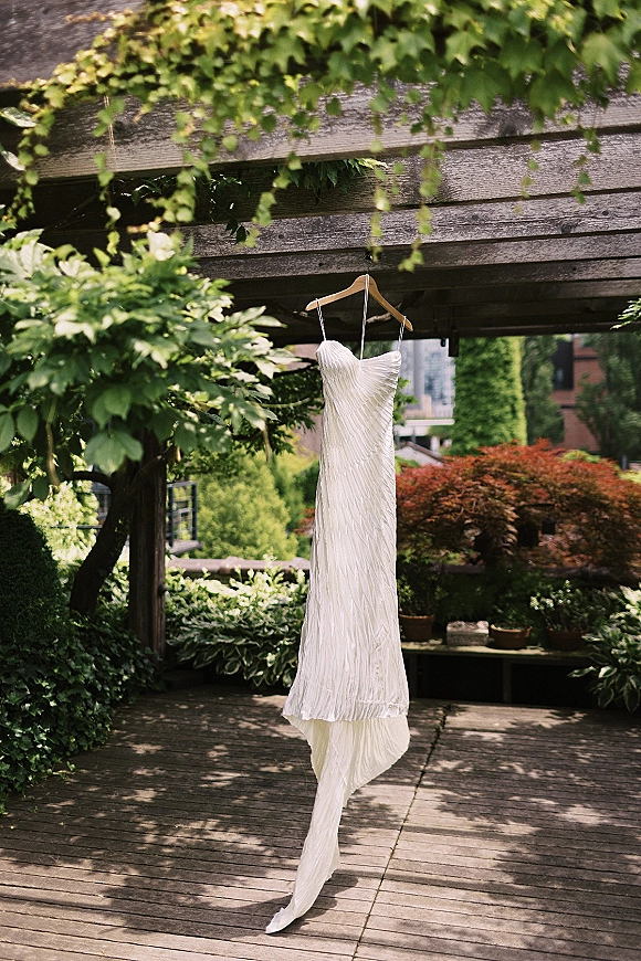 Wedding dress hanging from a wooden hanger, strapless white bridal gown displayed under a wood pergola with lush garden greenery behind