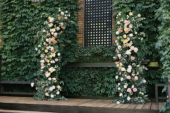 Wedding ceremony backdrop with floral ceremony pillars of blush, ivory, and yellow roses plus eucalyptus, set against an ivy-and-brick wall lattice