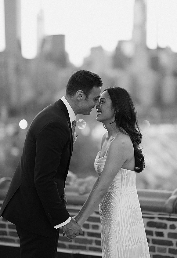 Couple portrait of bride and groom in a wedding kiss portrait, holding hands on a rooftop by a brick wall with city skyline behind