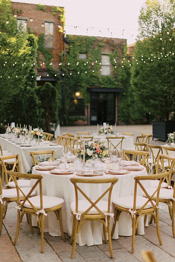 Outdoor reception decor with round tables in white linens, cross back chairs, floral centerpieces, taper candles under string lights in ivy brick courtyard