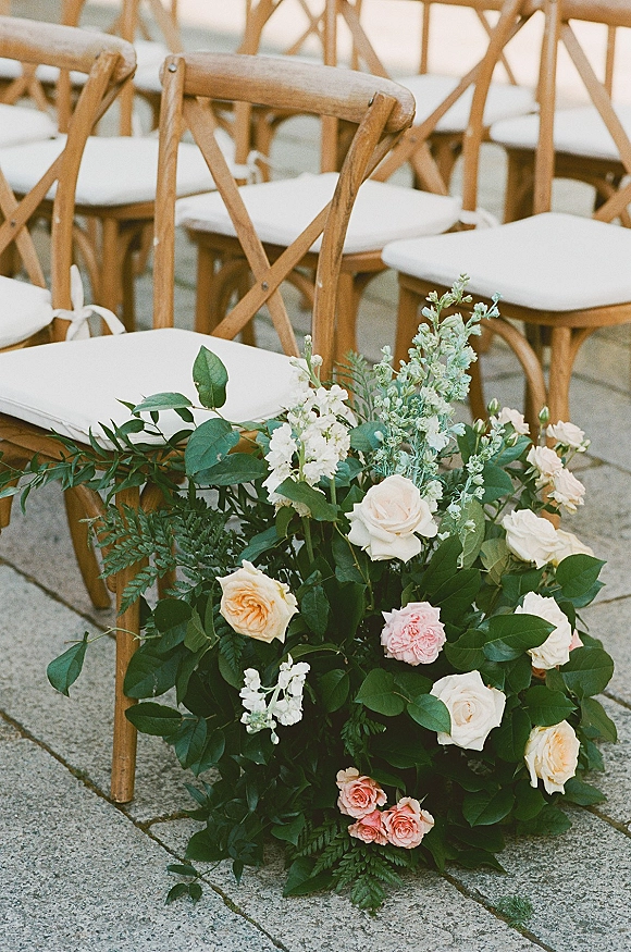 Ceremony aisle decor with wedding aisle flowers, a ground rose and fern arrangement beside white-cushioned crossback chairs on a stone patio