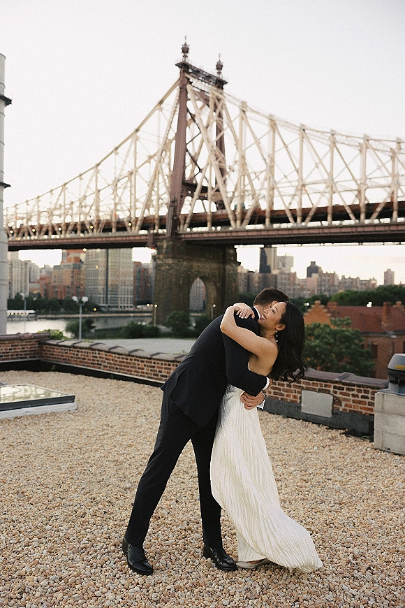 Couple portrait of bride in a strapless wedding dress and groom in a black suit embracing on a rooftop with bridge and city skyline at dusk